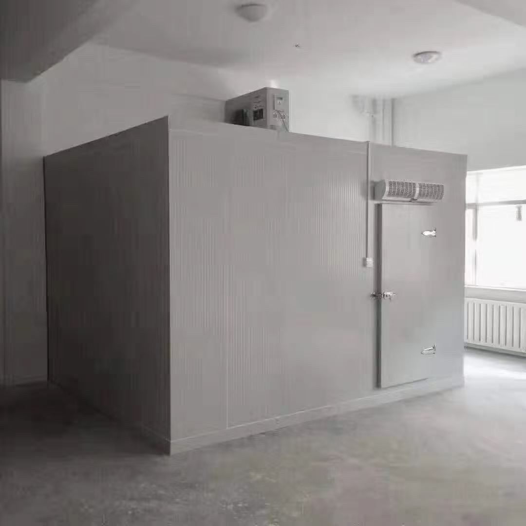 A compact panel room stands inside a larger indoor space. The front has a single hinged door, with equipment visible above the enclosure.